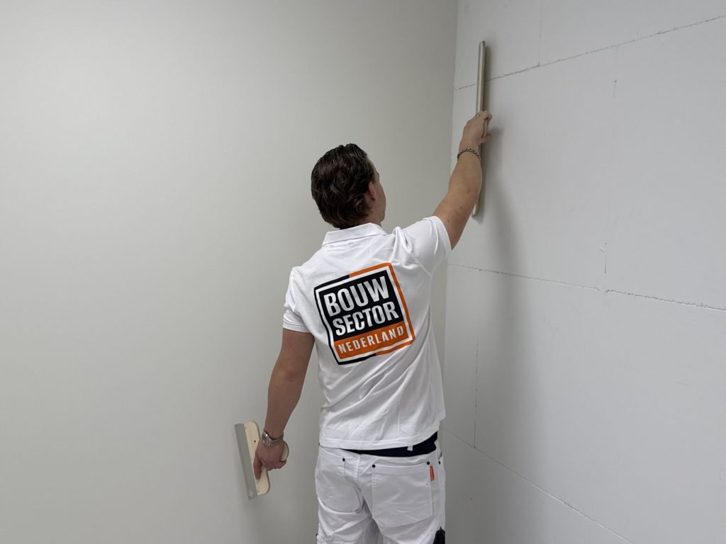 Stukadoor met Bouwsector-logo op zijn rug trekt met een lange spaan een wand naast de binnenhoek vlak